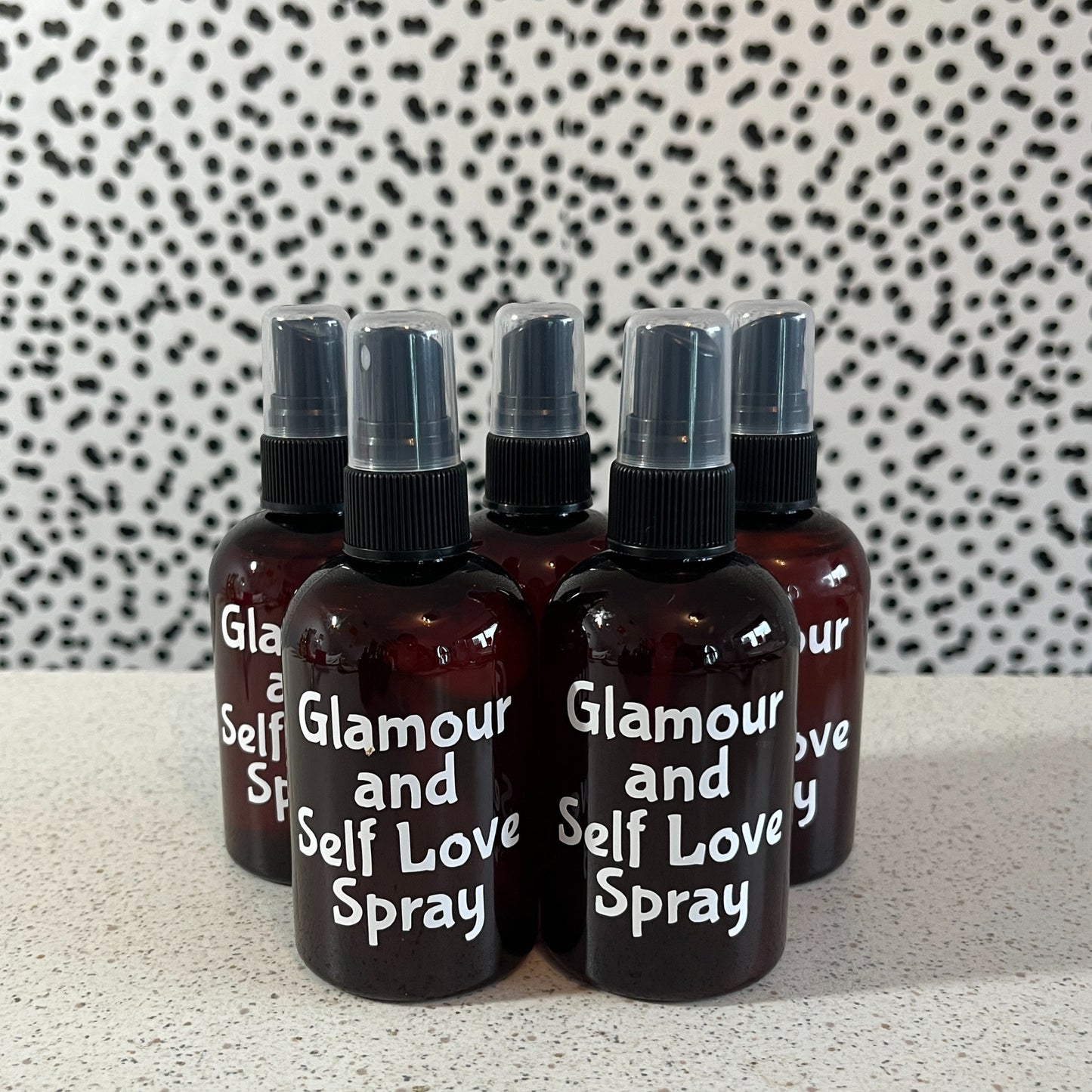 Set of 'Glamour and Self Love Spray' bottles on a speckled surface with a patterned background.