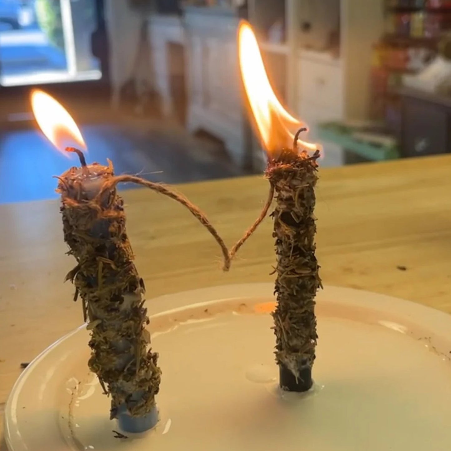 Two lit candles with natural wicks on a white plate against a blurred indoor background